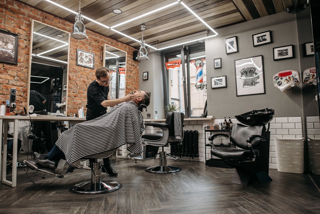A barber attends to a client in a stylish, contemporary barbershop with brick walls and framed art.